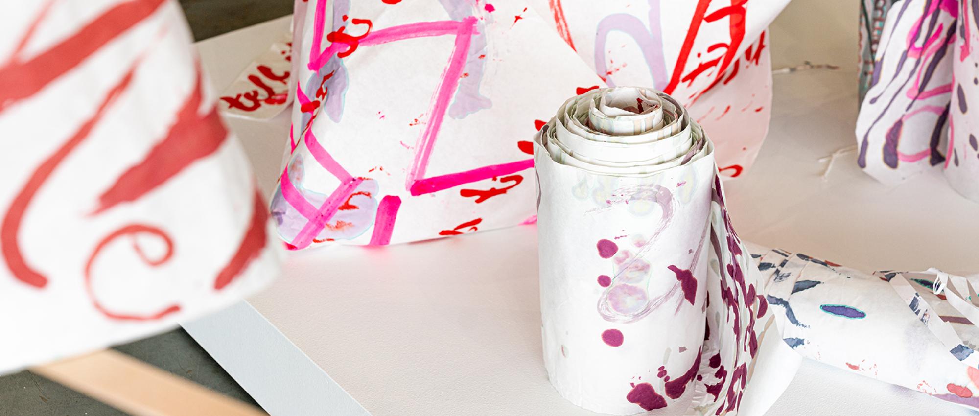 Paper scrolls sit upright on a low white pedestal. One hangs in the foreground. The scrolls bear text in reds and pinks that a curious viewer could decipher in person. From the photo you can make out an "e" here, a "W" there. Photo credit: Kevin Lopez.