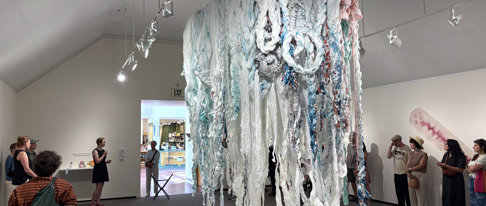 A white woman wearing a black dress holds a small microphone and speaks to a group of people standing around the edges of a hanging sculpture. The piece is mainly light blue and white, made of spun and twisted paper.